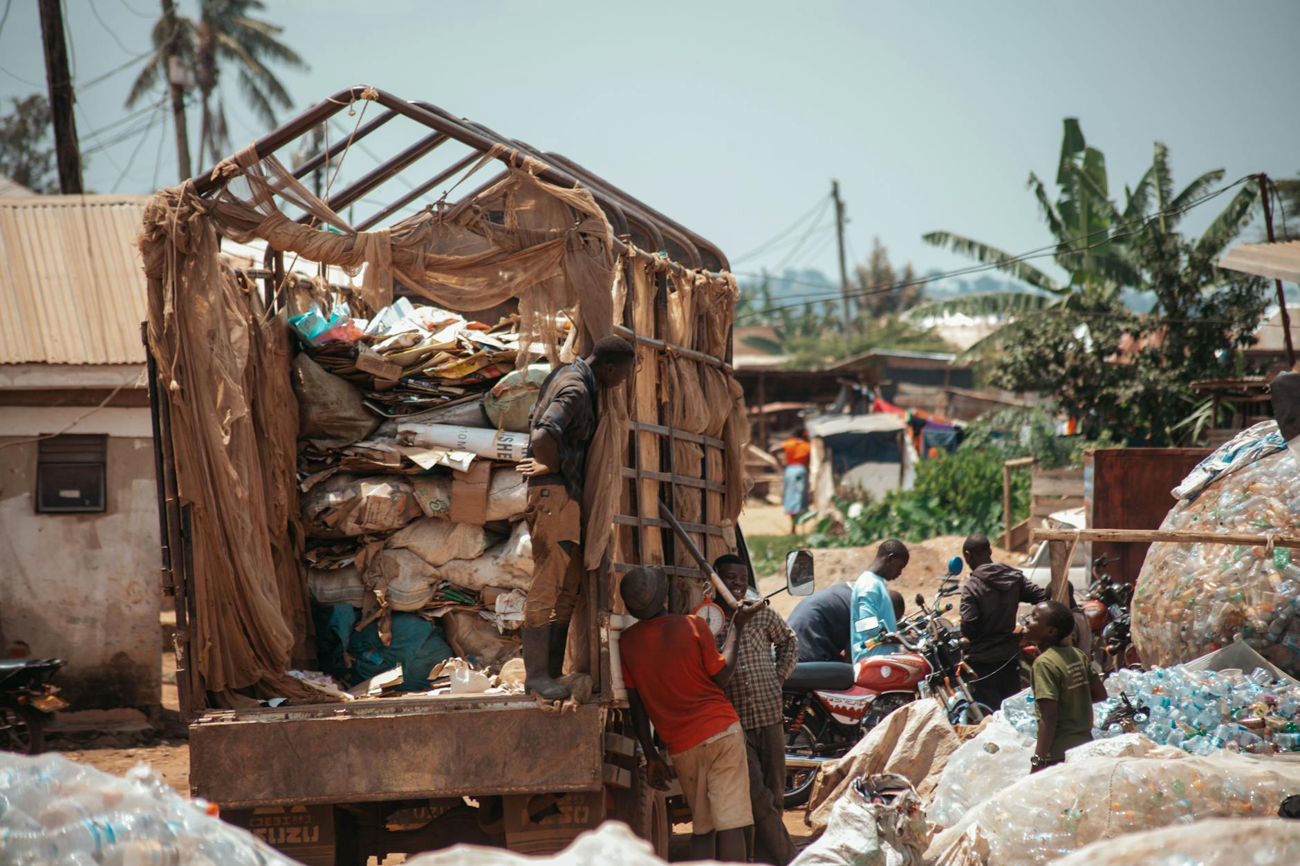 group of people in a garbage sorting plant next to bags of plastic bottles and a truck with waste paper